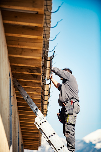 Roofer builder worker finishing folding gutter. He is up a ladder, photo taken from ground looking up, low angle view. He wears a tool belt, sky and clouds